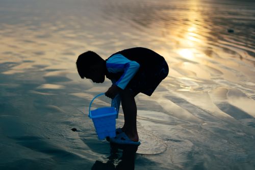 Free Young boy with a blue bucket exploring beach sands during a scenic sunset. Stock Photo