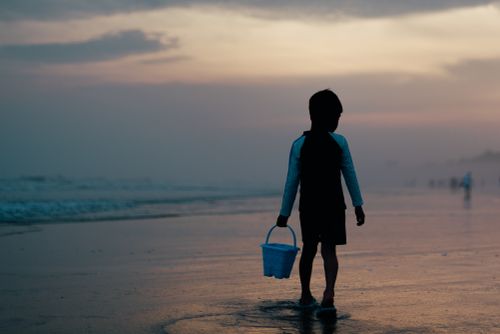 Free A child carries a bucket while walking along a beach at sunset, creating a serene silhouette scene. Stock Photo
