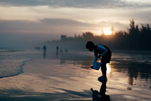 Free A child playing with a bucket on the beach at sunrise, casting a silhouette against the calm sea. Stock Photo