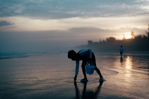 Free Child with a bucket bending over on a misty beach during sunset, capturing a serene moment of exploration. Stock Photo