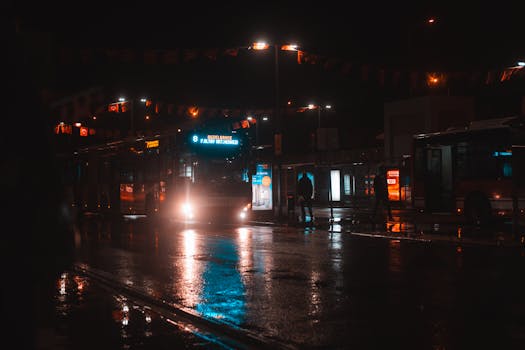 A city bus illuminated in the night at a busy station with wet streets.