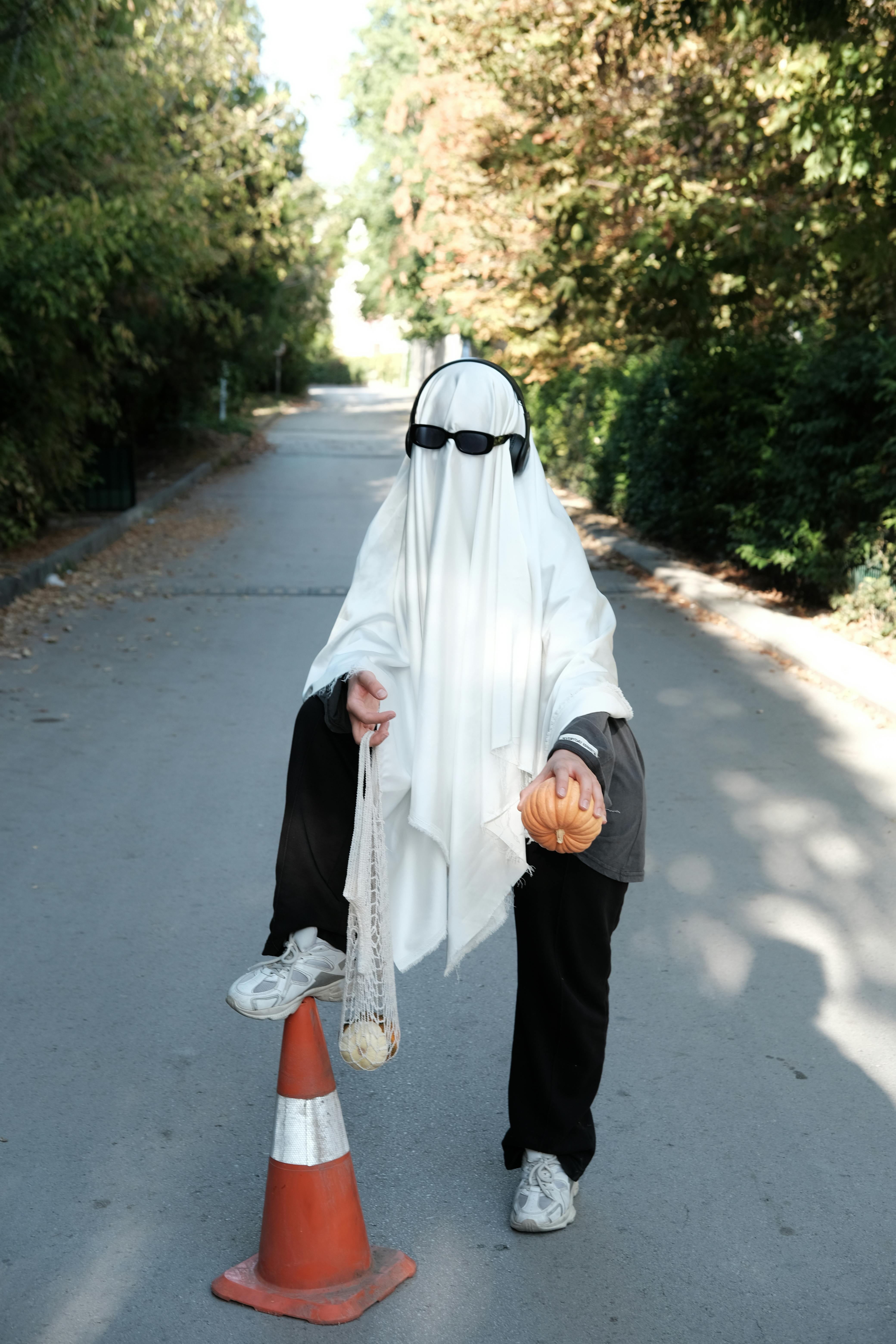 Person in ghost costume with pumpkin and headphones posing on an urban street for Halloween.