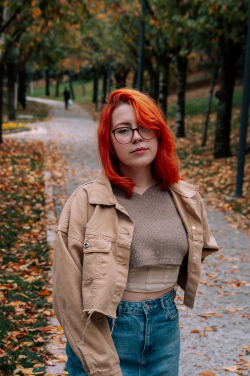 Free Young woman with red hair poses stylishly in a fall park, wearing a casual outfit and eyeglasses. Stock Photo