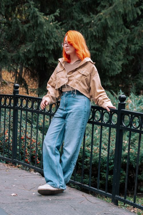 Free Stylish woman with red hair in casual attire posing against a fence in a park. Stock Photo