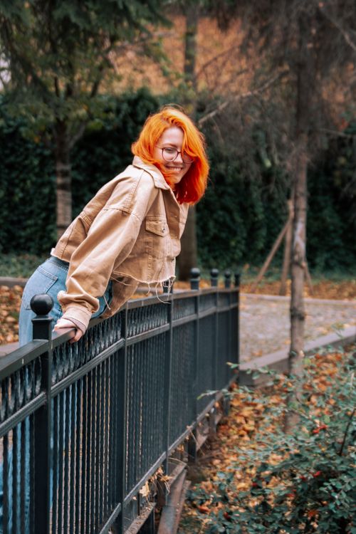 Free Redheaded woman leaning on a park fence, enjoying a bright autumn day with vibrant colors. Stock Photo