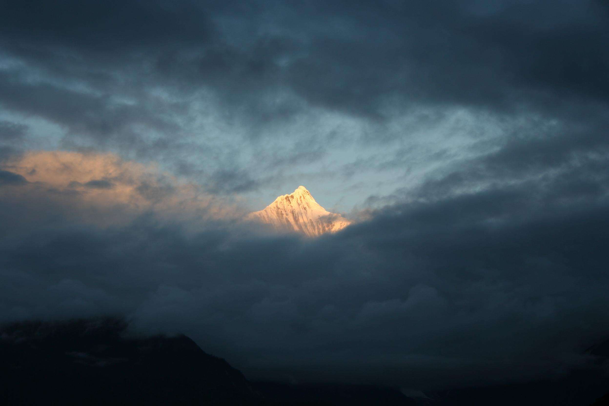 A dramatic scene of a mountain peak emerging through clouds, lit by warm sunlight, creating a striking contrast.