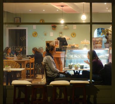 Two women engaged in conversation inside a cozy cafe at night, seen through a window.