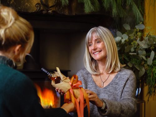 Free A joyful moment of gift-giving between two women by a cozy fireplace, surrounded by greenery. Stock Photo
