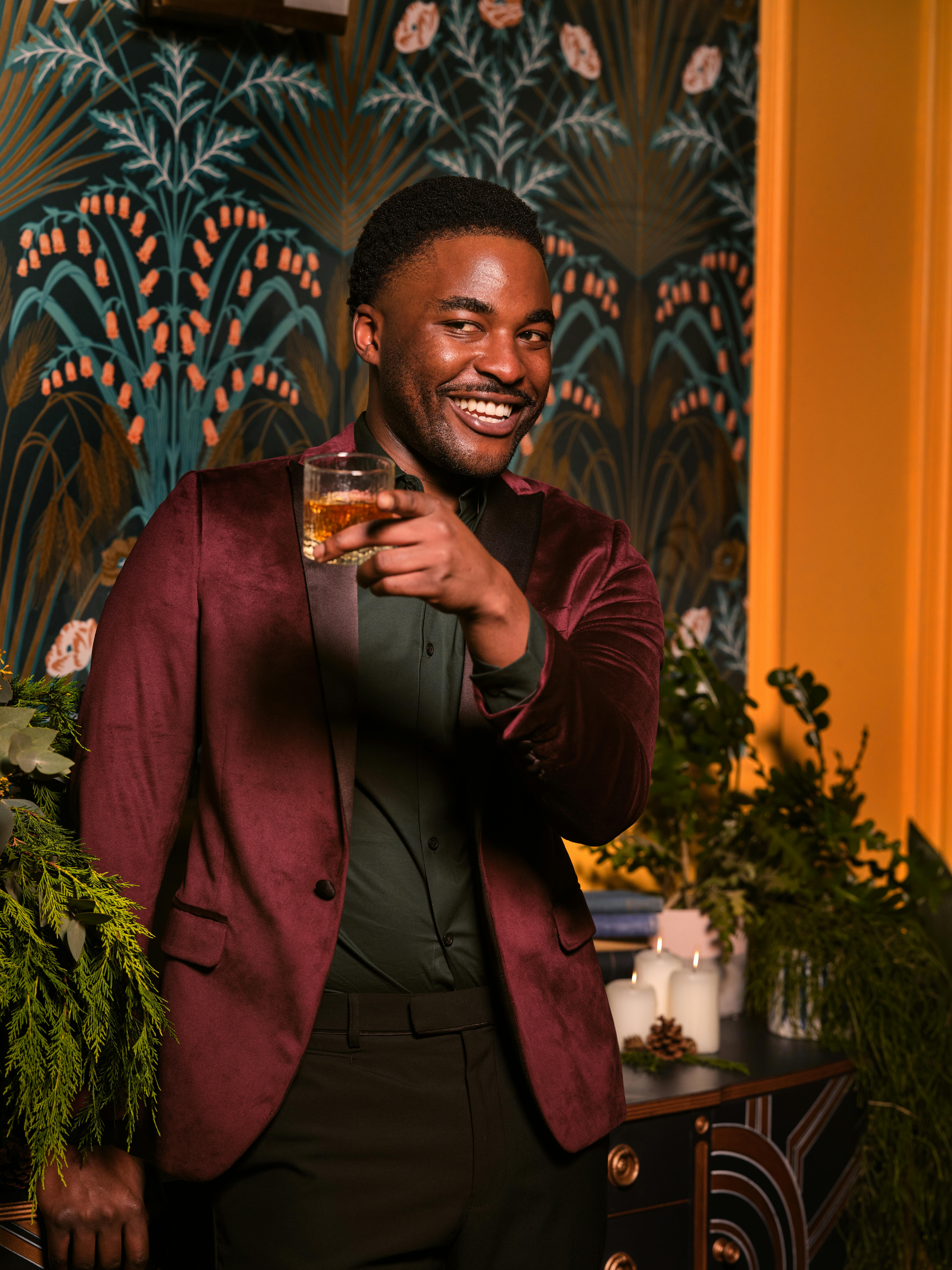 Man in burgundy suit holding a drink, smiling at a festive indoor setting with floral wallpaper.