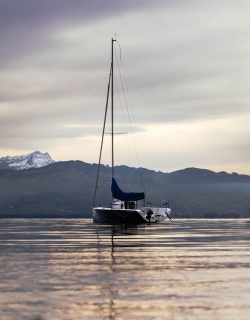 Free A serene view of a sailboat on Bodensee, surrounded by mountains and reflected in calm waters. Stock Photo
