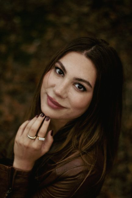 Close-up portrait of a smiling woman with long brunette hair wearing a brown jacket outdoors.