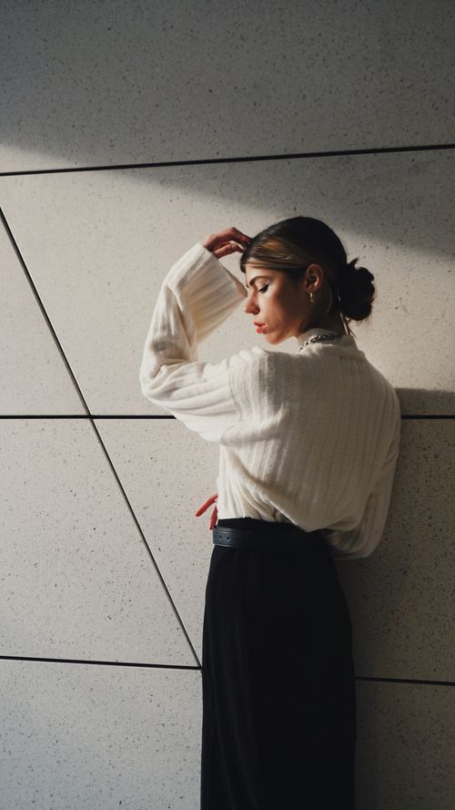 Free Young woman in chic white sweater and black pants posing against a contemporary wall design. Stock Photo