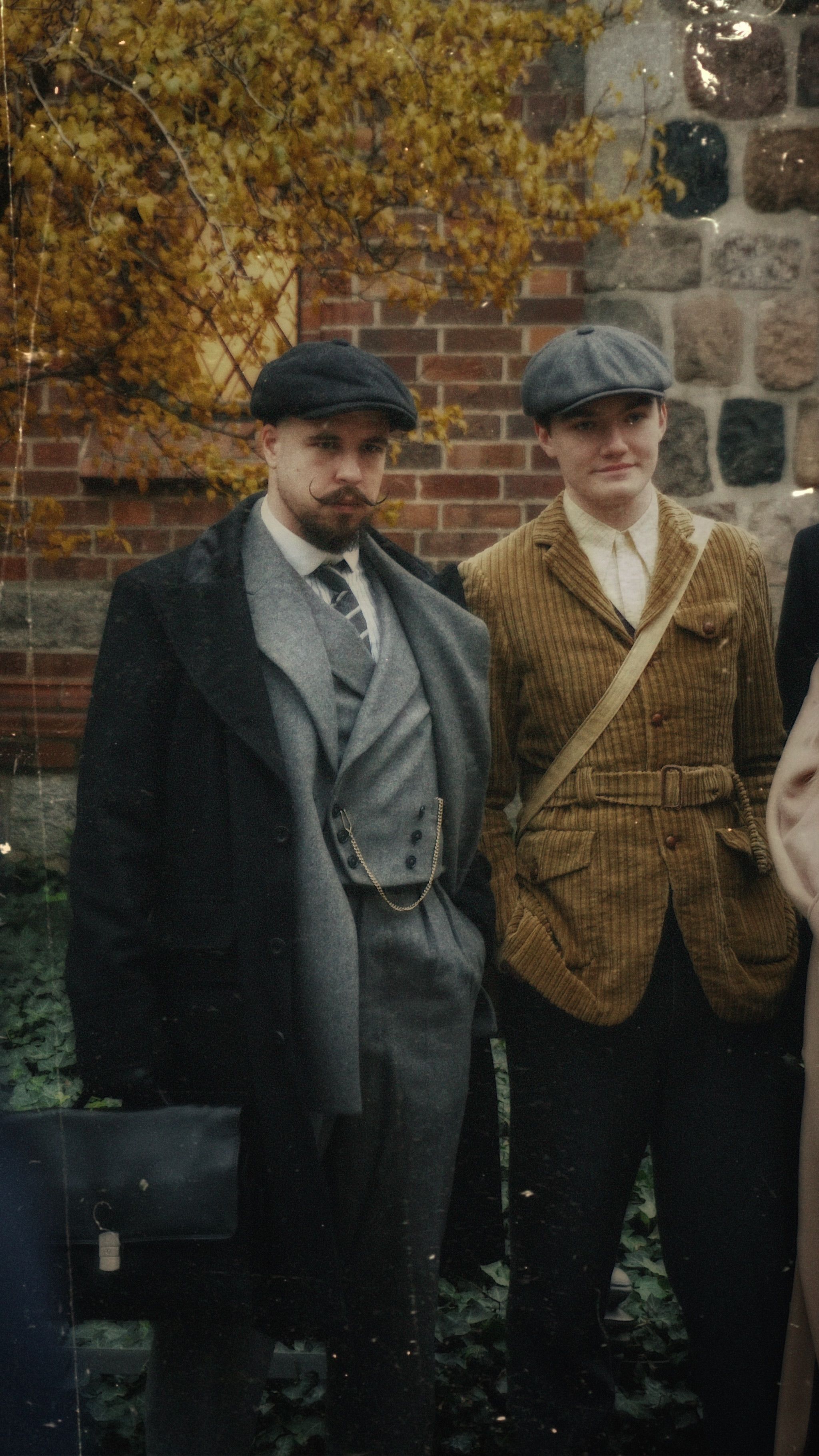 Two men in vintage suits and caps posing outdoors against a brick wall, capturing a nostalgic retro style.