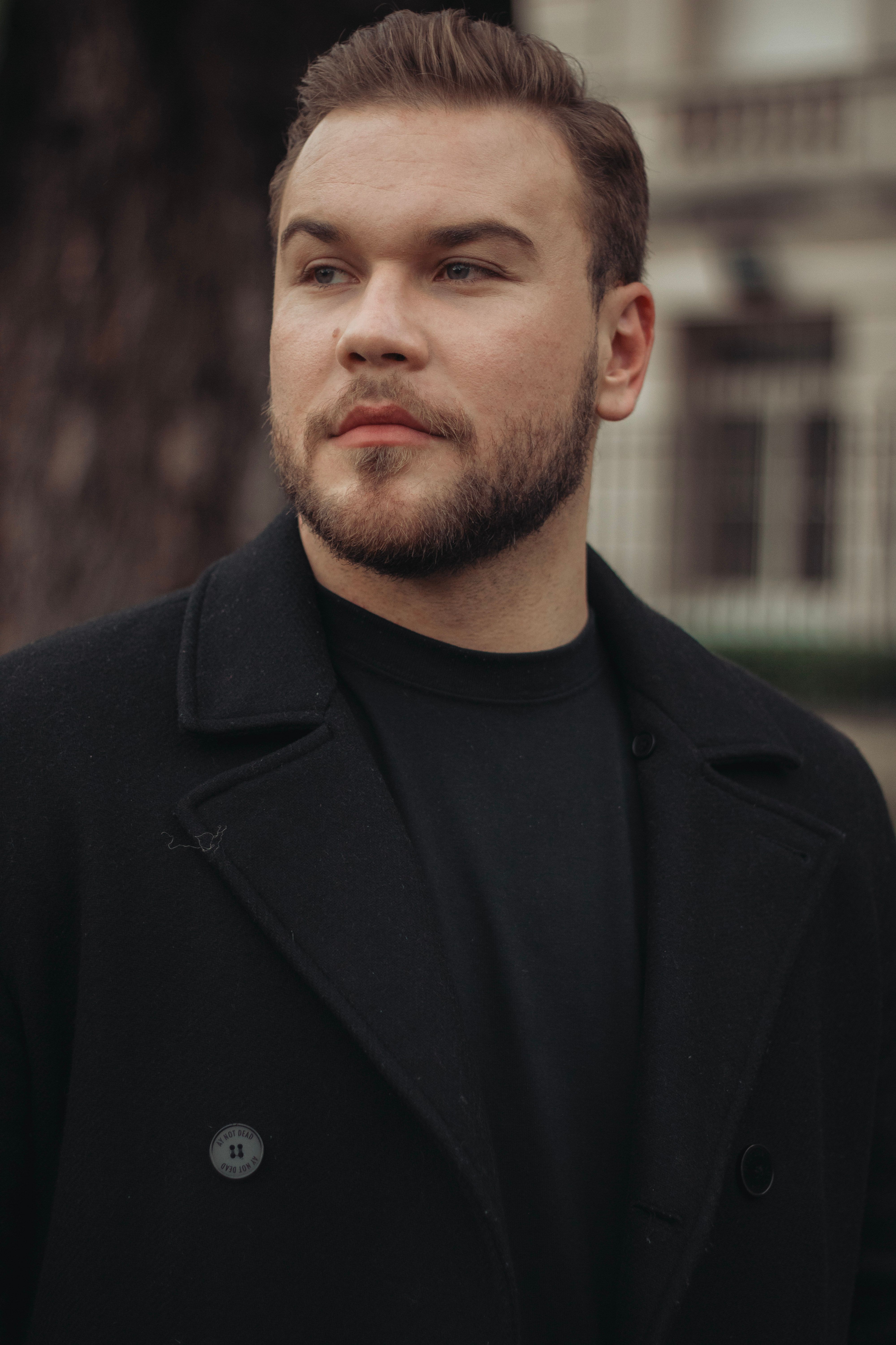 A trendy man with a beard poses in a city setting wearing a fashionable jacket.