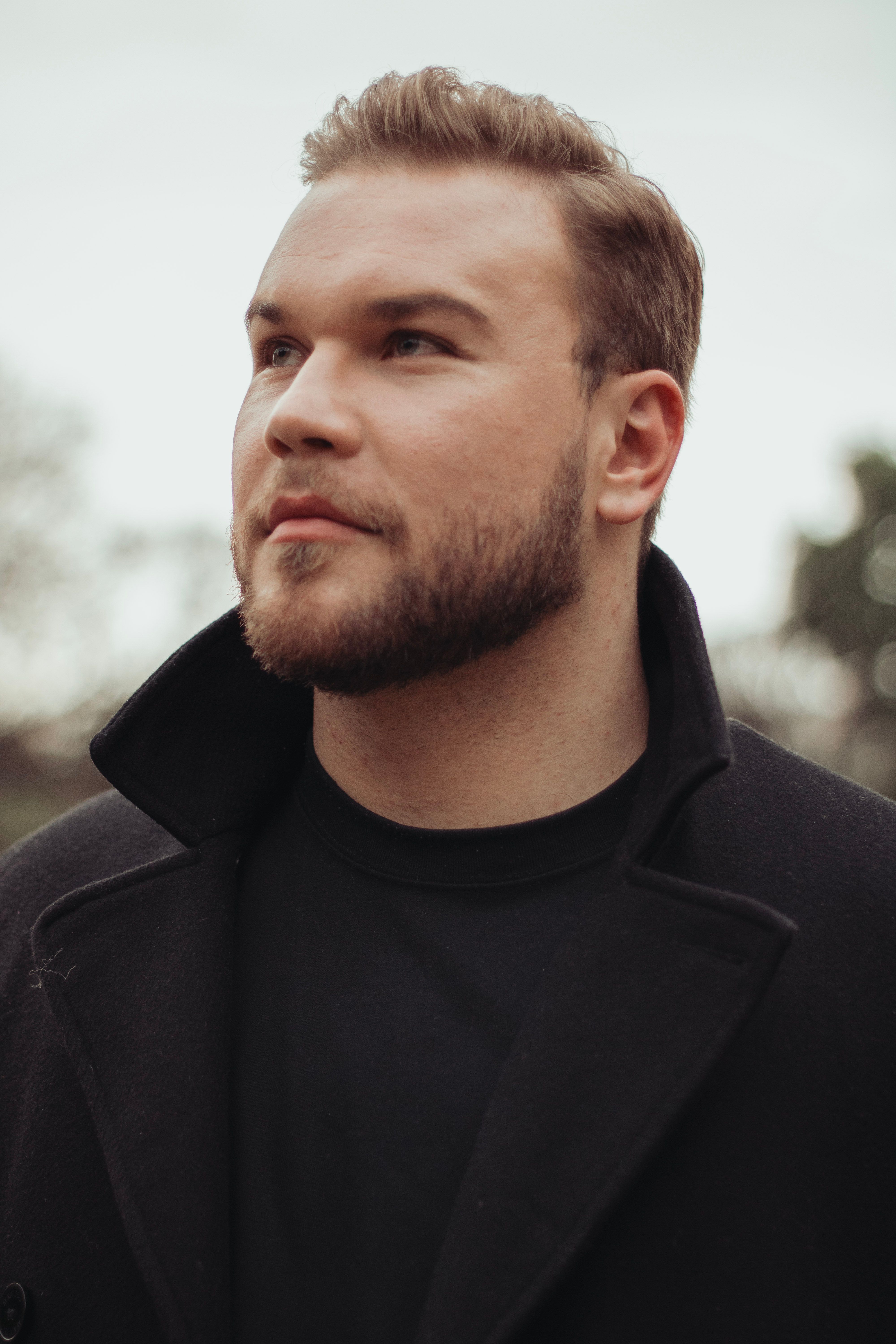 Man with a beard in a black coat looks upwards outdoors. Casual fashion portrait.
