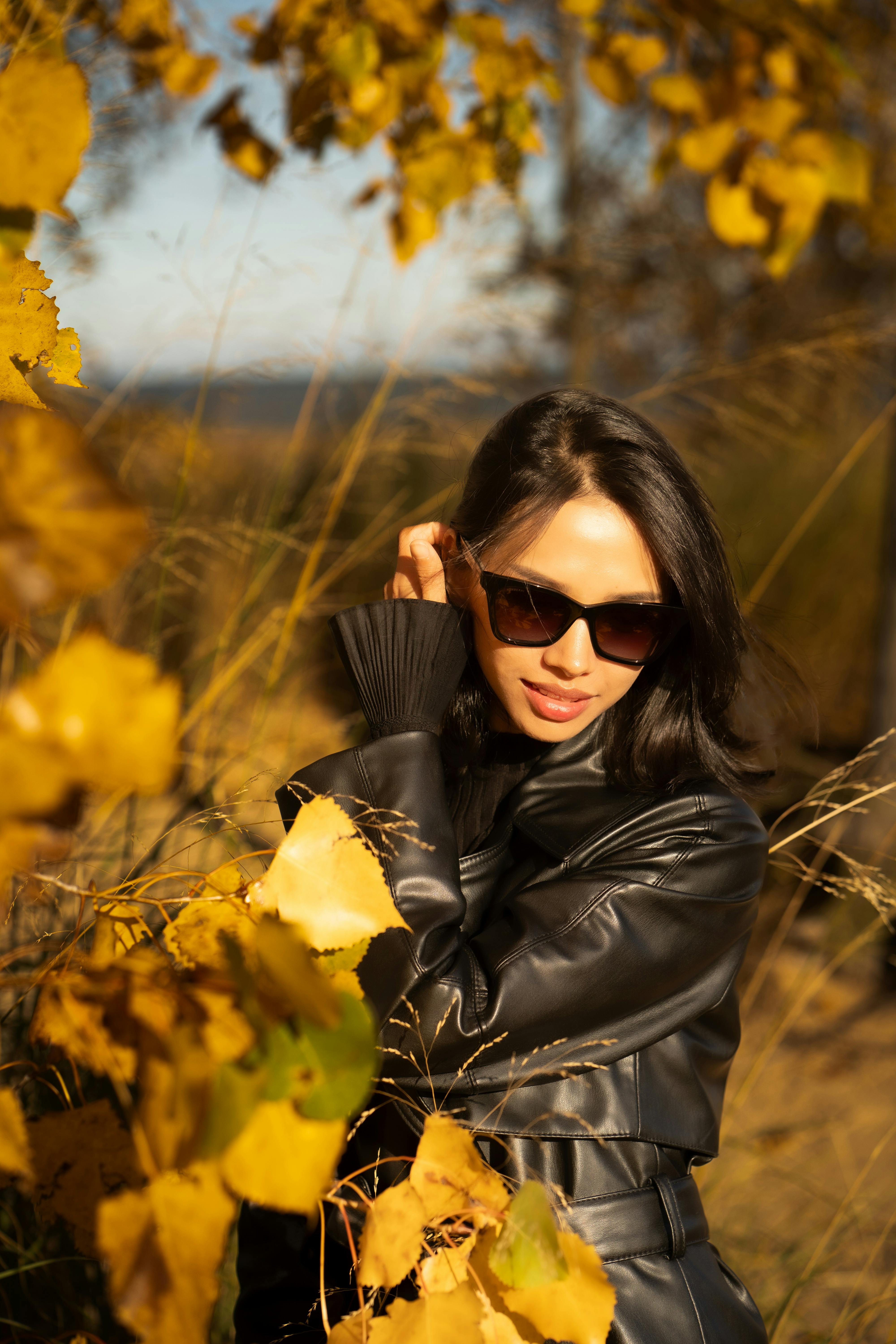 Fashion portrait of a woman in sunglasses and leather jacket surrounded by autumn foliage during the day.