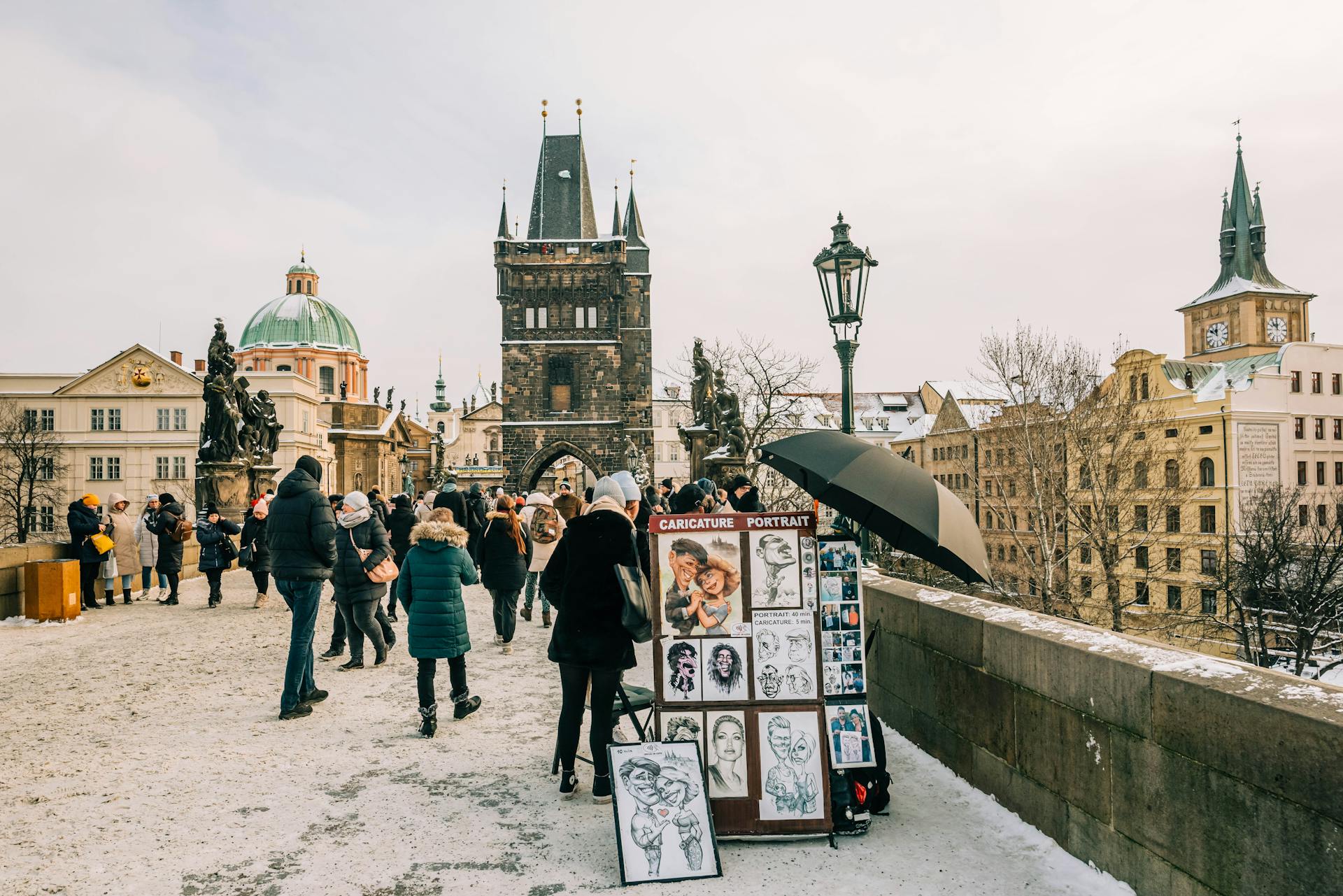 People walking on the snowy Charles Bridge in Prague, depicting local landmarks and street art.