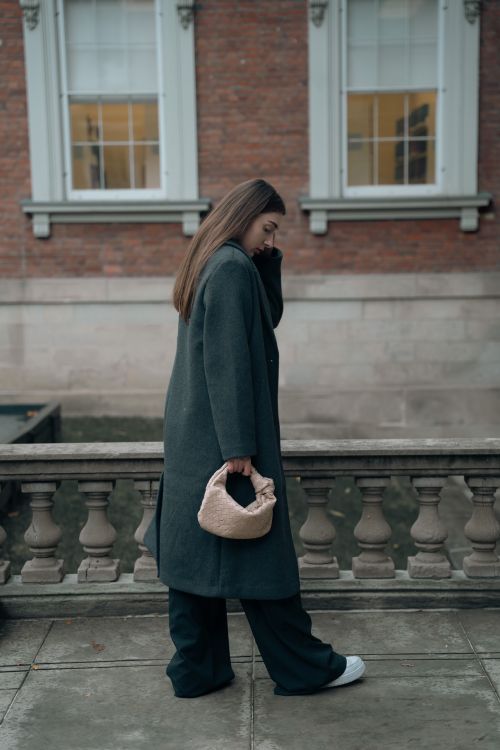 Free Stylish woman in a long coat walking outdoors, holding a small bag. Stock Photo