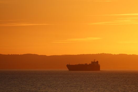 A cargo ship silhouette against a dramatic orange sunset over calm waters.
