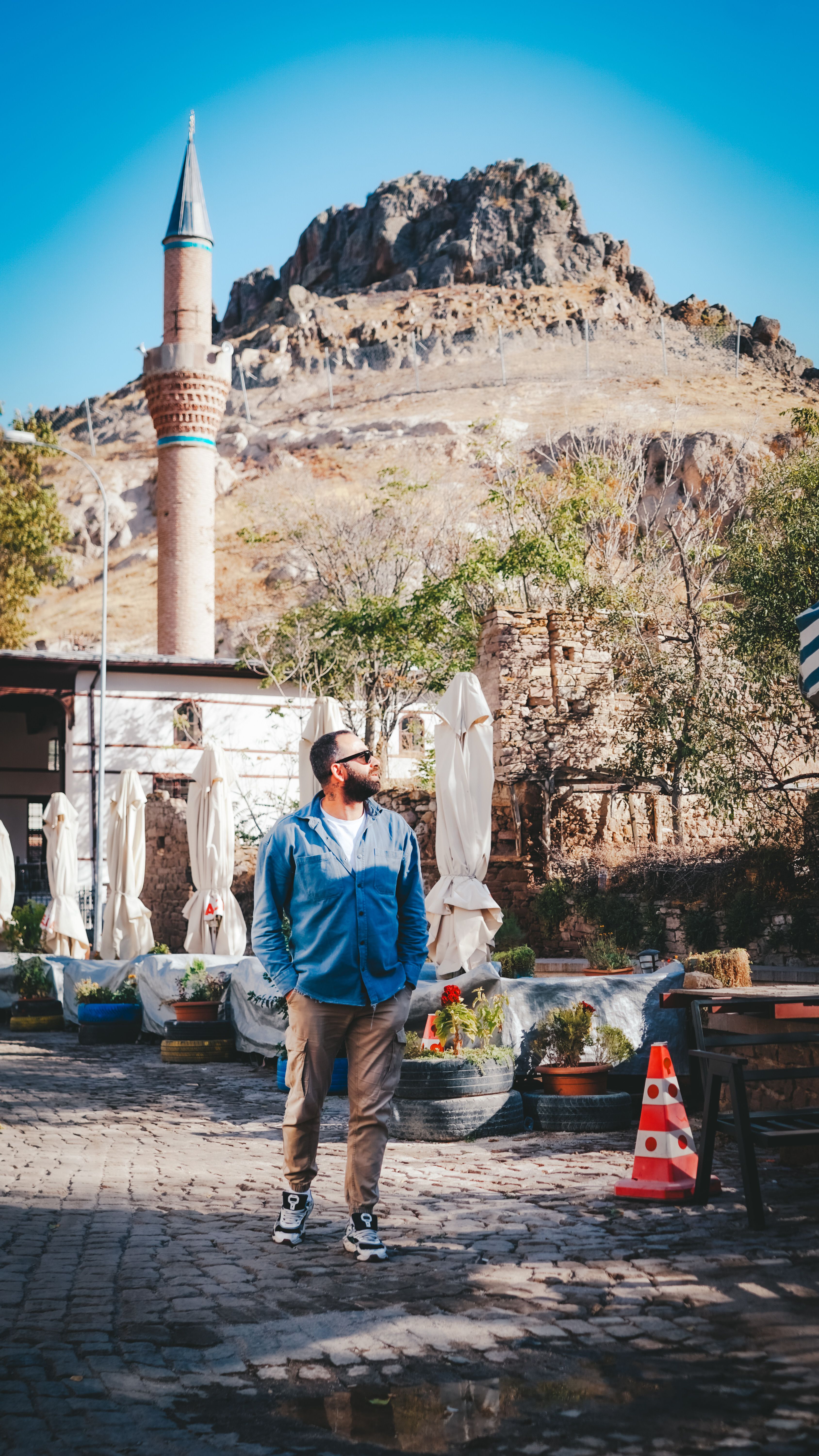 A man stands in an outdoor courtyard with a historic minaret and mountain backdrop.