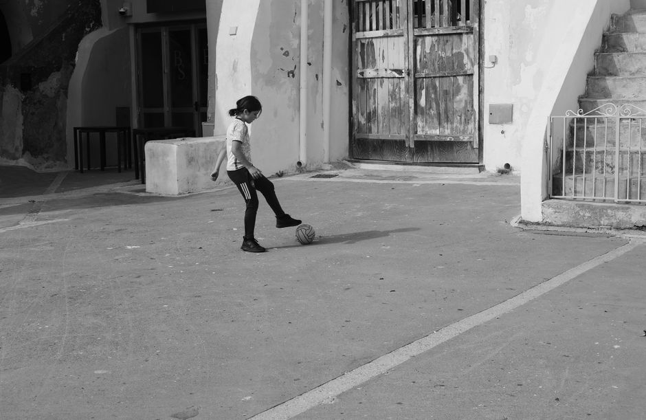 A young girl plays soccer alone on a quiet urban street in black and white.