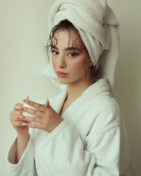 Elegant woman in bathrobe with a towel wrapped around her hair holding a cup.