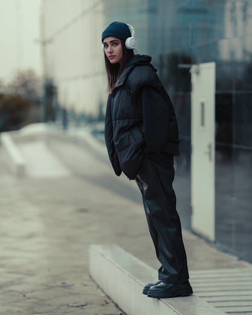 Young Woman in a Black Fashionable Outfit and Headphones Standing on a Sidewalk in City 