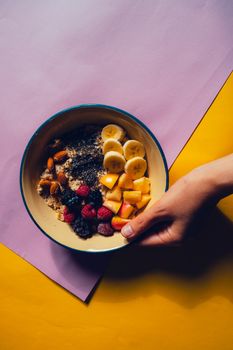Hand holding a vibrant fruit bowl with bananas, chia seeds, and berries on a colorful background.