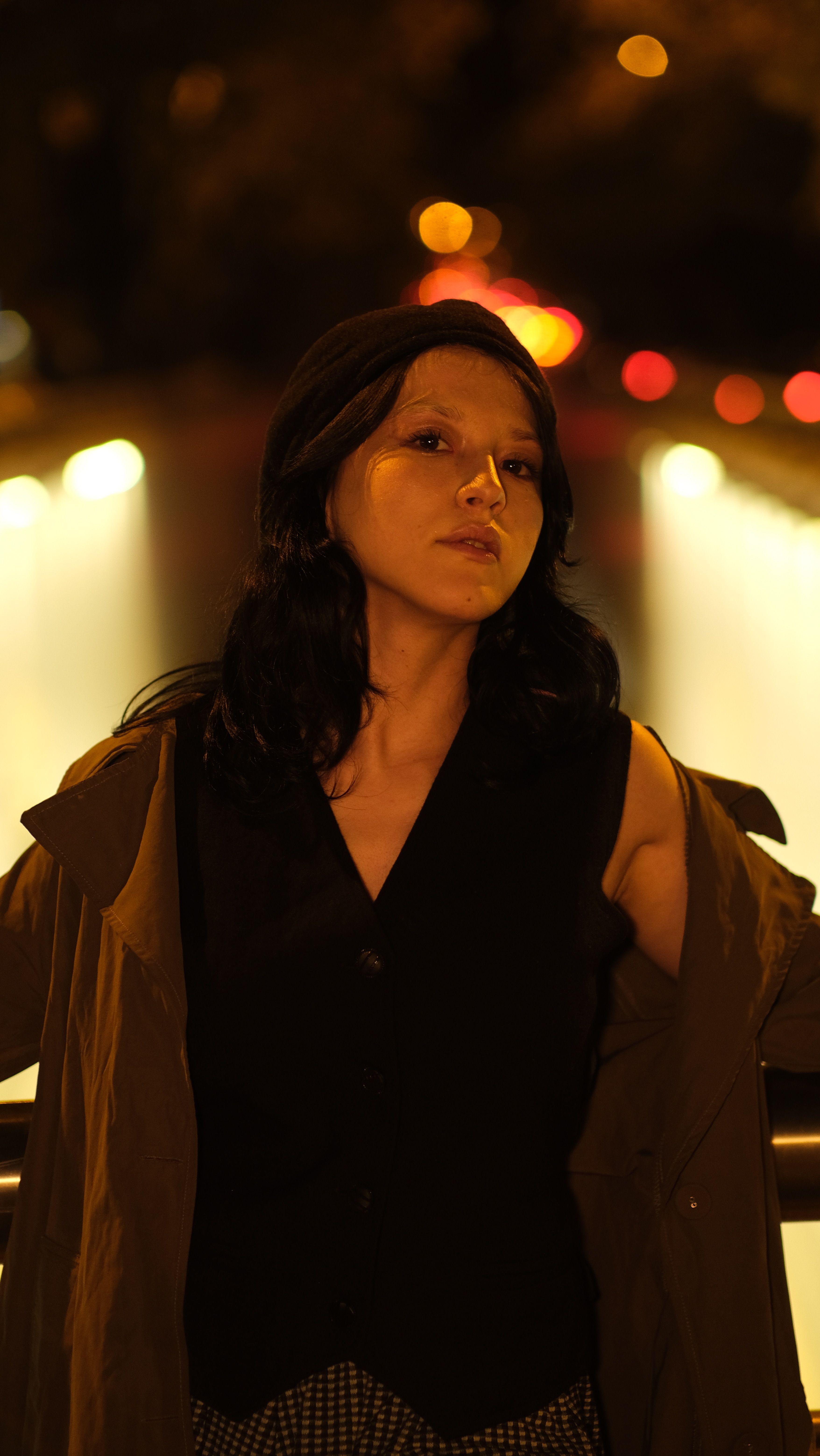 A fashionable woman posing at night with city lights in the background, wearing a coat and looking serene yet serious.