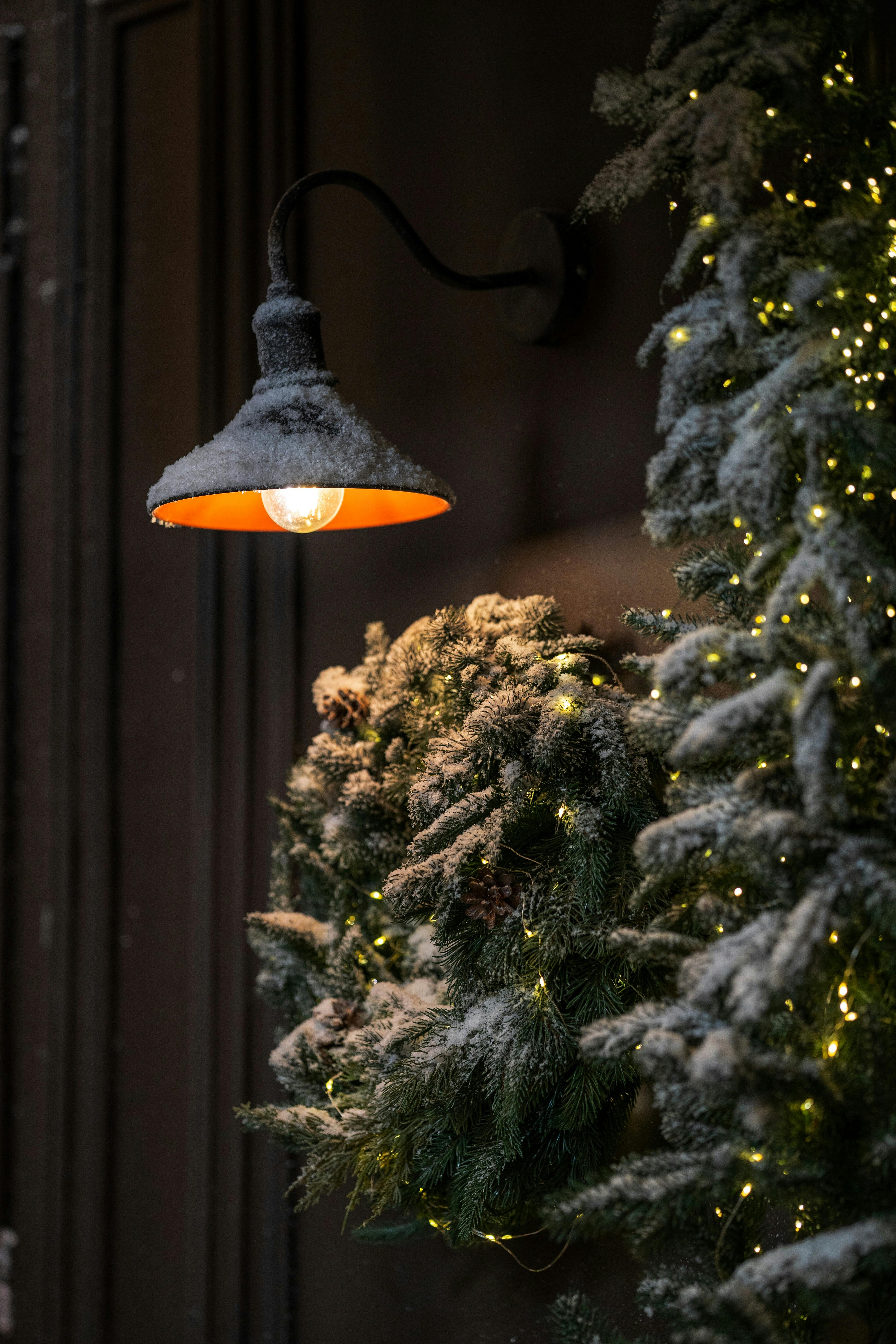 A wintery night scene featuring a snow-covered lamp and decorated greenery with fairy lights.