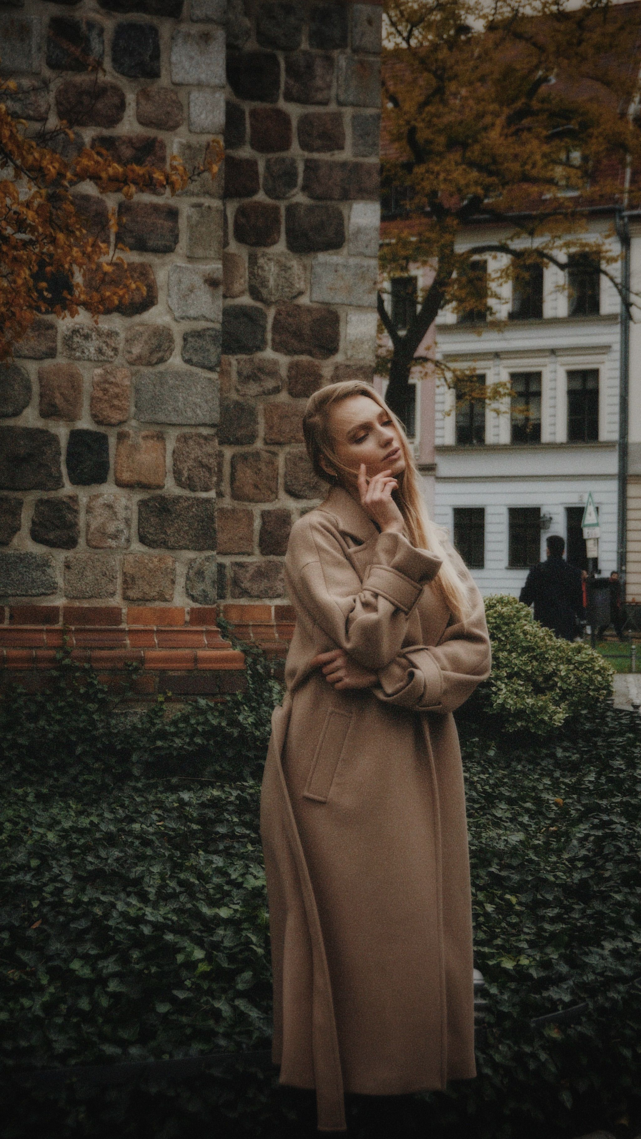 Fashionable woman in a coat standing thoughtfully against a stone wall in an urban setting.