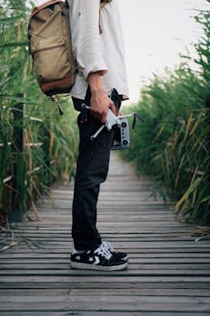 A man stands on a wooden boardwalk surrounded by reeds, holding a drone, symbolizing adventure and exploration.