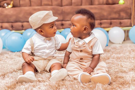 Two cute babies sitting on a rug surrounded by blue and white balloons indoors.