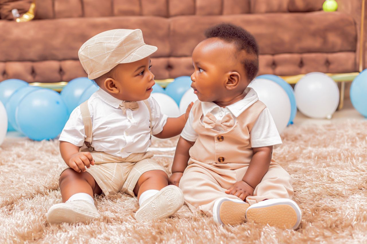 two cute babies sitting on a rug surrounded by blue and white balloons