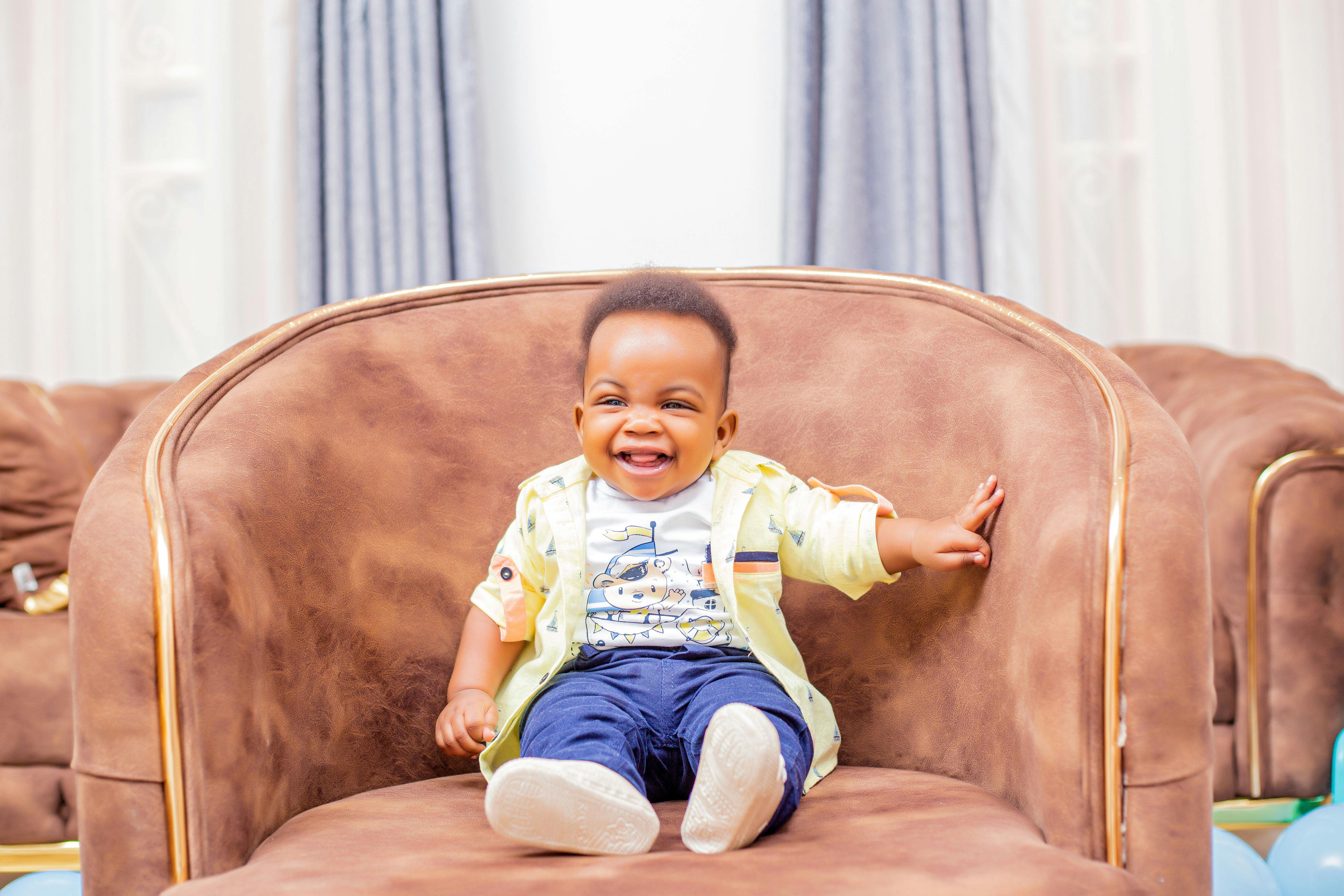 Charming baby boy laughing while sitting in a brown armchair indoors. Perfect for family and lifestyle themes.