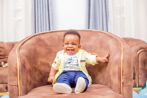 Free Charming baby boy laughing while sitting in a brown armchair indoors. Perfect for family and lifestyle themes. Stock Photo