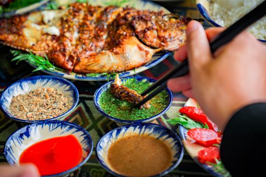 A vibrant display of Asian cuisine with grilled fish, dips, and a hand using chopsticks.