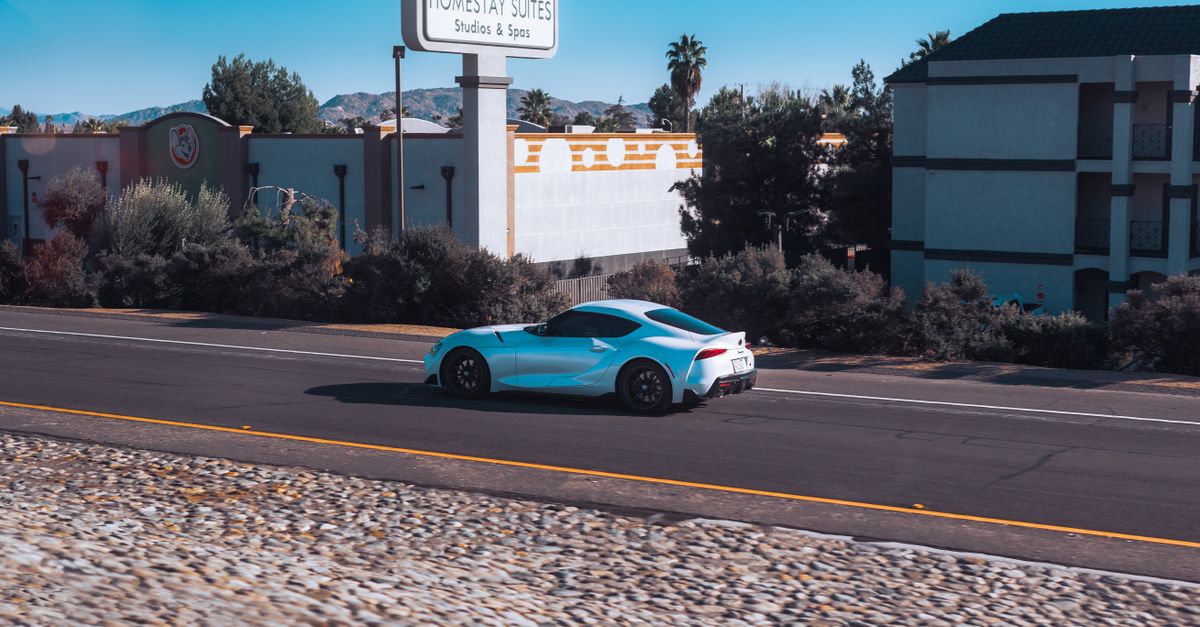 Blue Toyota GR Supra driving on road past Homestay Suites. Sunny day, clear sky.