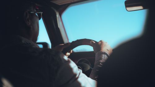 Free A man in a striped shirt driving a car with focus on steering wheel under clear blue sky. Stock Photo