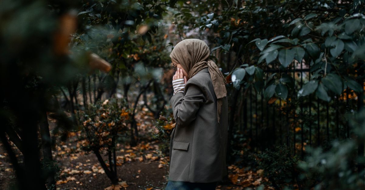 A woman wearing a hijab walks thoughtfully through a leafy autumn park.