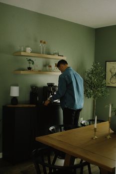 Adult male in jean jacket making coffee in a stylish home kitchen with wooden shelves.