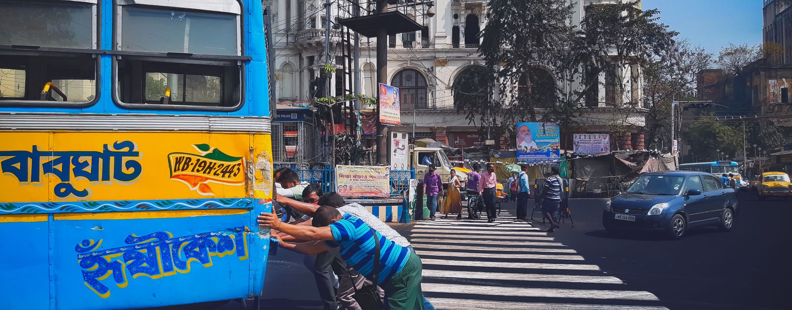 Street scene in Kolkata, India with people pushing a bus at a crosswalk in a busy urban setting.
