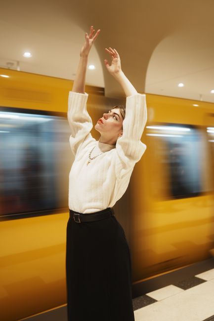 Young woman raises arms gracefully as a subway train passes by in a Berlin metro station.