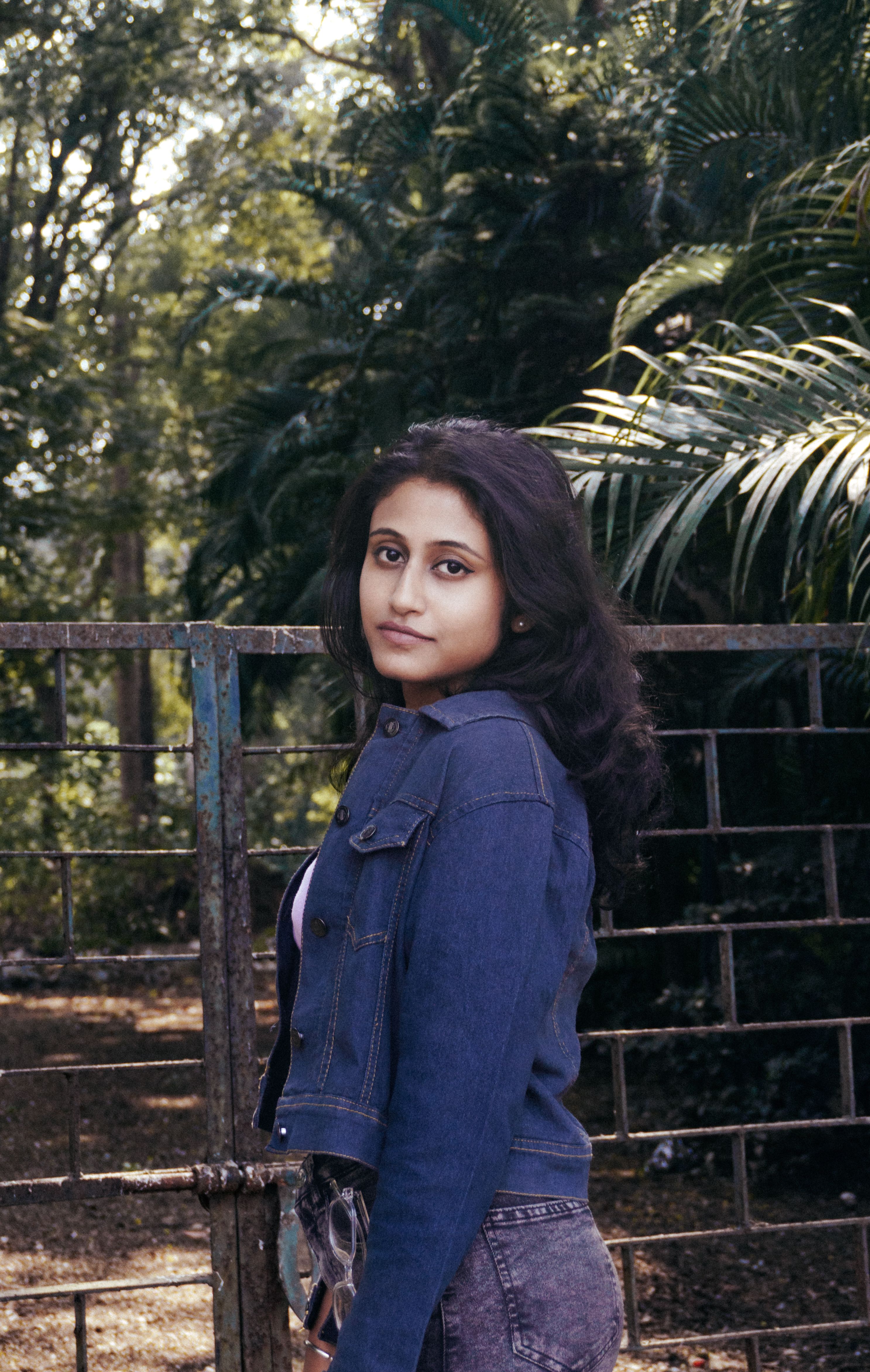 A young woman wearing a denim jacket stands confidently outdoors, surrounded by greenery.