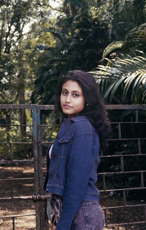 Free A young woman wearing a denim jacket stands confidently outdoors, surrounded by greenery. Stock Photo