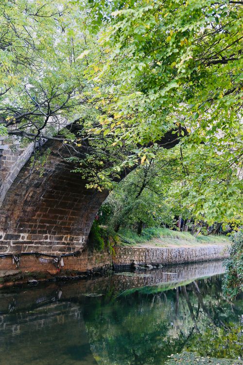Free A stone bridge arches over a tranquil river surrounded by lush greenery, offering a peaceful nature scene. Stock Photo