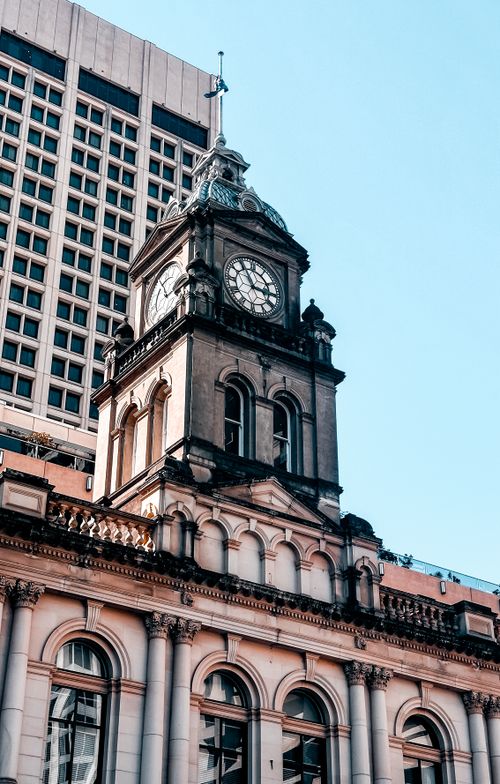 Free A vintage clock tower stands amidst modern urban architecture under a clear sky. Stock Photo