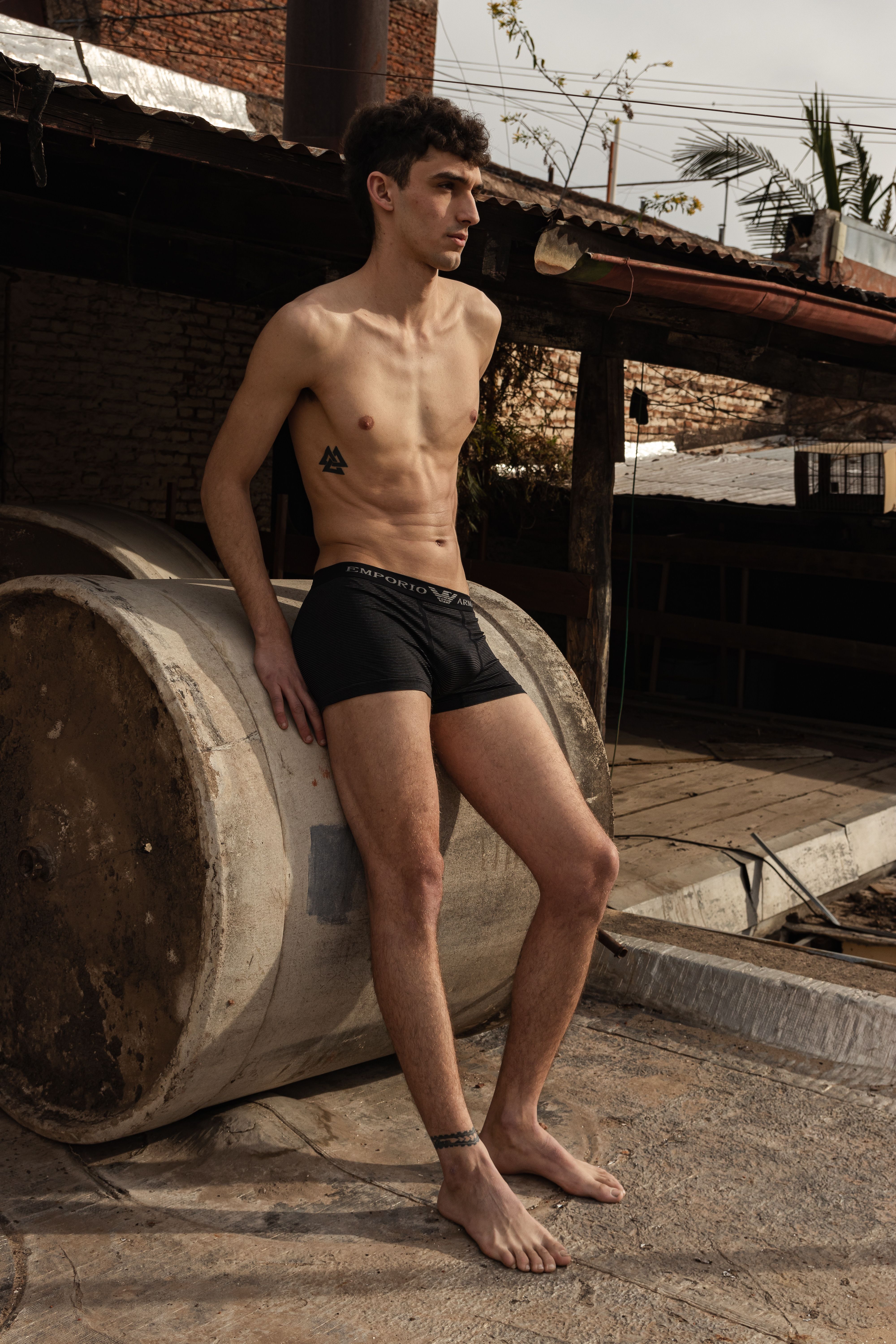 A shirtless male model in boxer shorts poses outdoors against a rustic backdrop, showcasing tattoos.