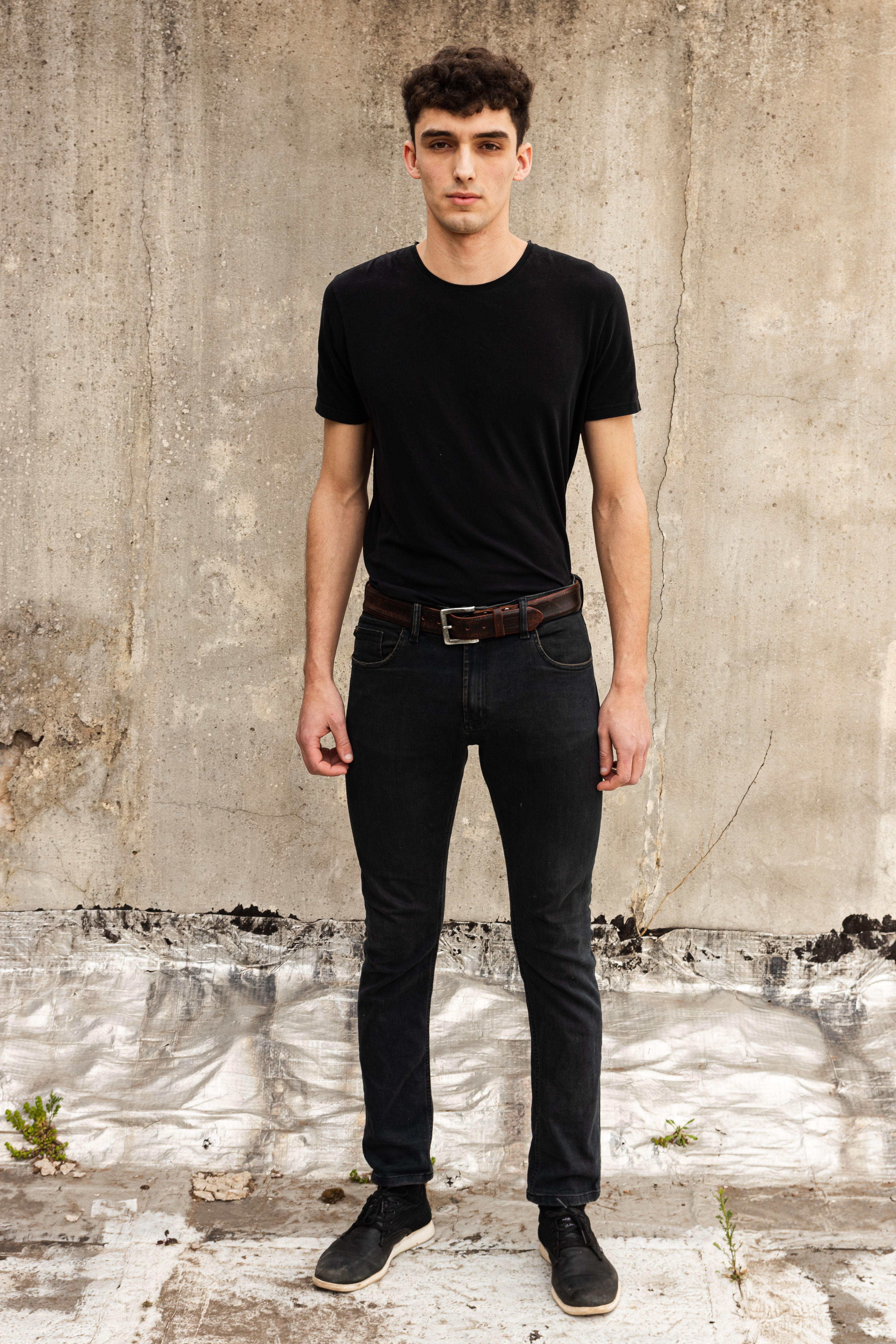 Casual fashion portrait of a man in black attire, standing against a textured urban wall.