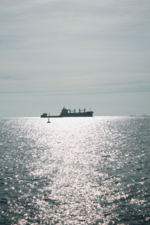 Free A solitary cargo ship sails across a sparkling ocean under a vast sky, creating a serene seascape. Stock Photo